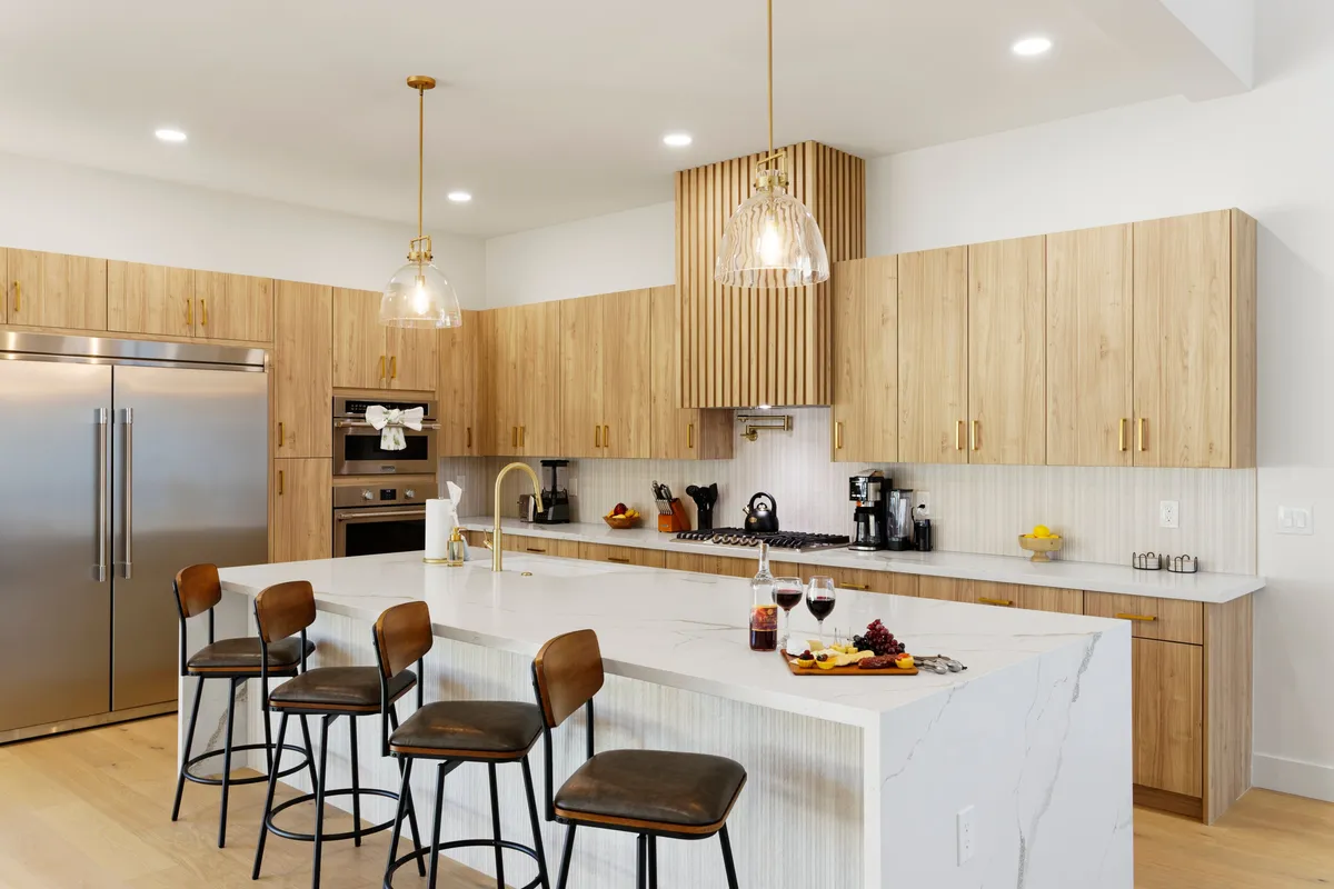 A stylish, open-concept kitchen with a large island surrounded by bar stools, gleaming stainless steel appliances, wood cabinetry, and elegant lighting fixtures. The space is clearly designed for both serious cooking and entertaining guests.