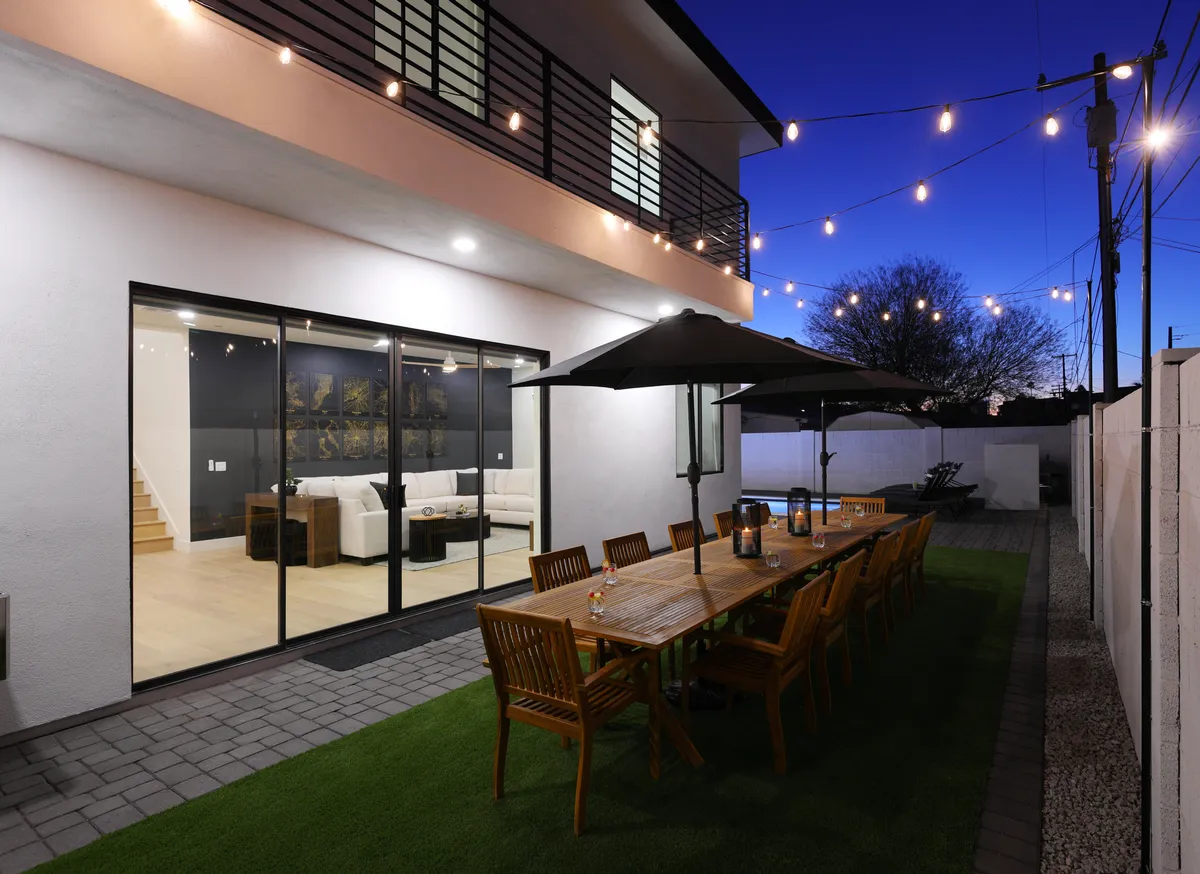 An inviting outdoor dining table sits beneath a canopy of string lights, with sliding glass doors providing seamless indoor-outdoor flow. The lounge area inside is visible, connecting the backyard’s vibrant social space with the home’s interior.