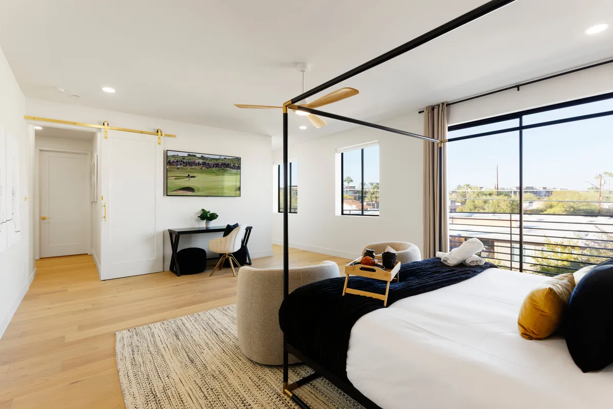 A spacious primary bedroom featuring a stylish four-poster bed, large windows letting in soft daylight, a desk/workspace area, and tasteful artwork. The neutral tones and uncluttered design evoke a sense of calm and comfort. The room’s layout subtly incorporates elements like the desk and plenty of closet space, reflecting the luxury and functionality described in the section.