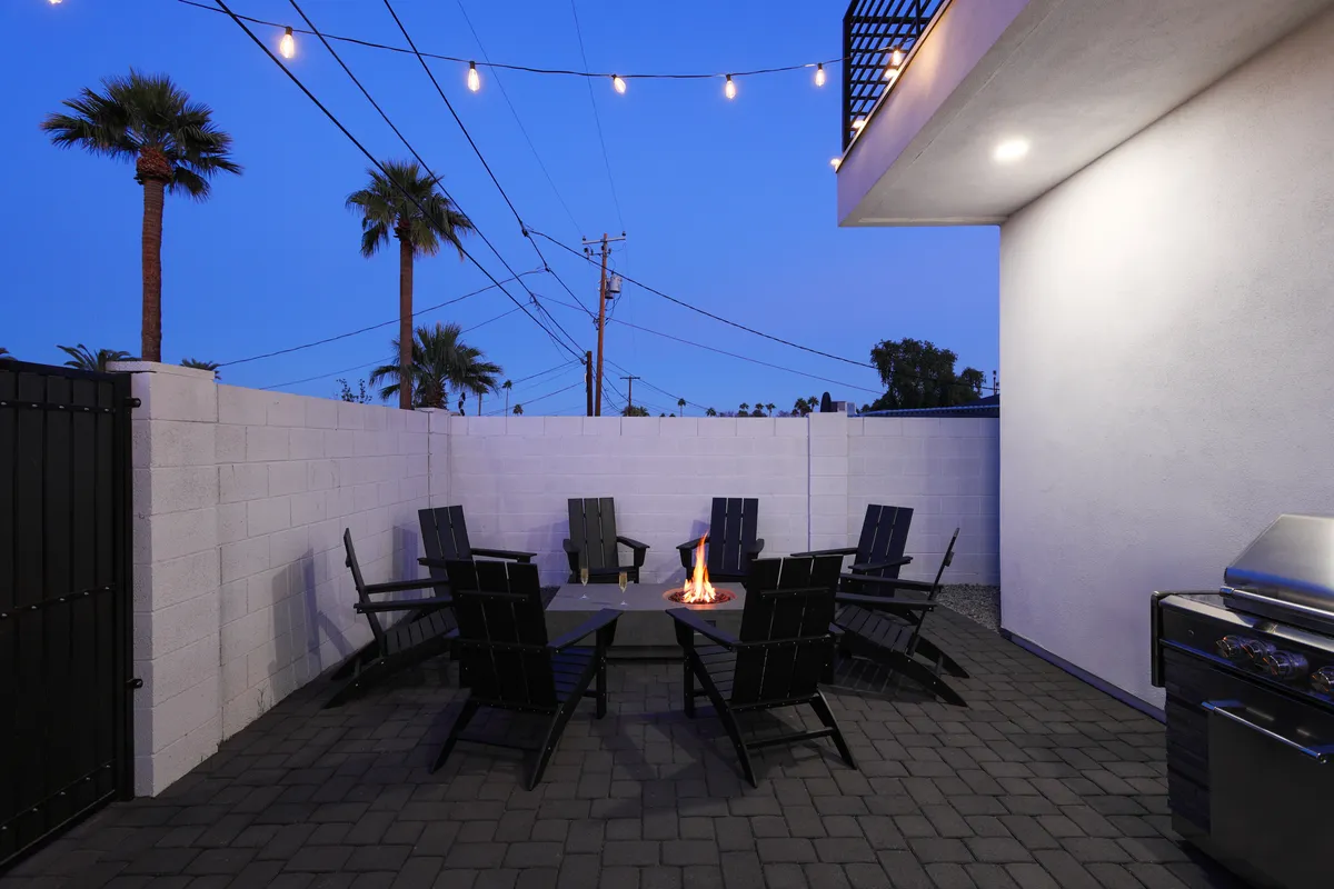 A cozy fire pit with surrounding seating, illuminated by string lights, and a nearby grill creates a warm communal atmosphere. This setup is perfect for gathering around the fire in the evening, sharing stories, or grilling outdoors.