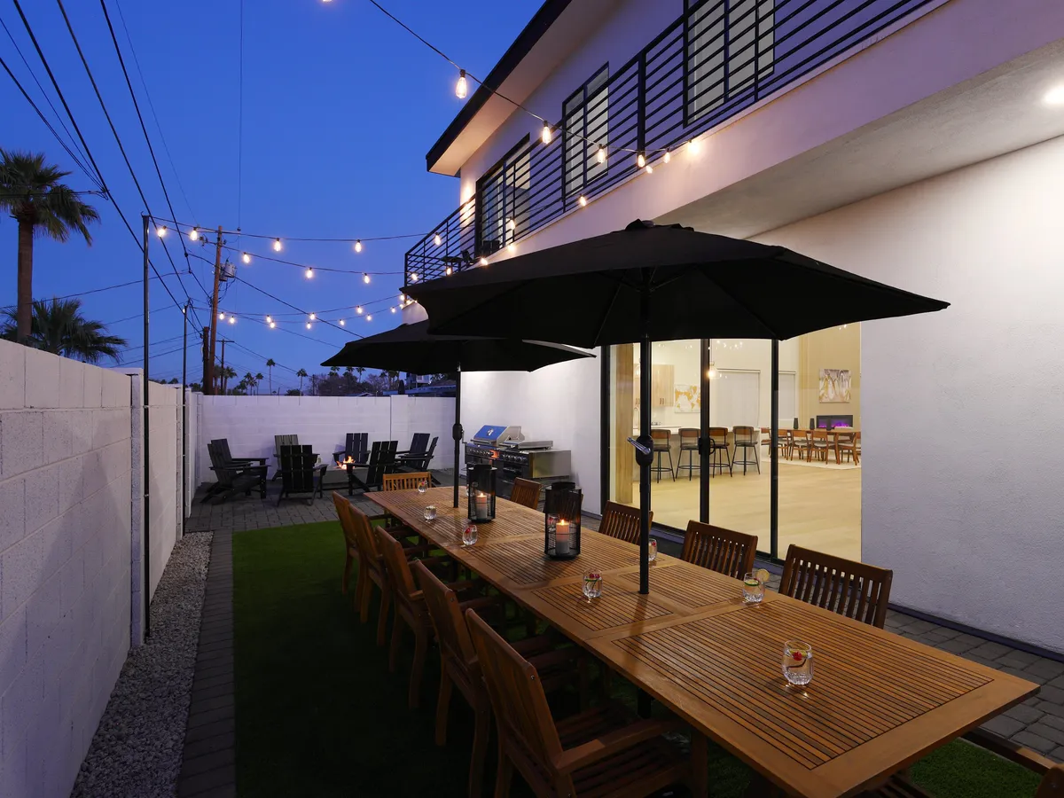 A festive outdoor dining area set beneath string lights, featuring a long wooden table, umbrellas for shade, and a fully equipped grill station. The setup invites group gatherings for al fresco meals as the sun sets, capturing the sociable and communal aspects of the backyard.