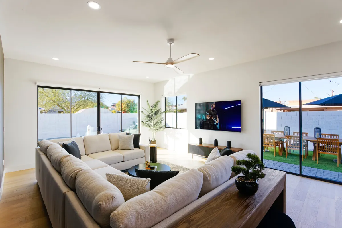 A spacious and inviting living room with a large sectional sofa facing a modern television, all bathed in natural light streaming through glass doors that open to an exterior view. The room exudes comfort and is ideal for relaxing or gathering with family and friends.