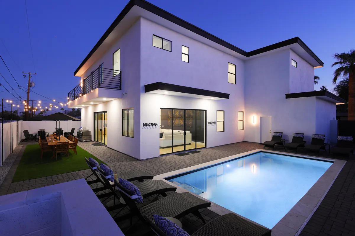 The image showcases a stylish modern home in Scottsdale with a sparkling swimming pool at center stage, surrounded by a chic outdoor dining area ideal for gatherings. Evening lighting casts a relaxed, festive glow, inviting guests to unwind, swim under the stars, or share meals with friends in a resort-like atmosphere.