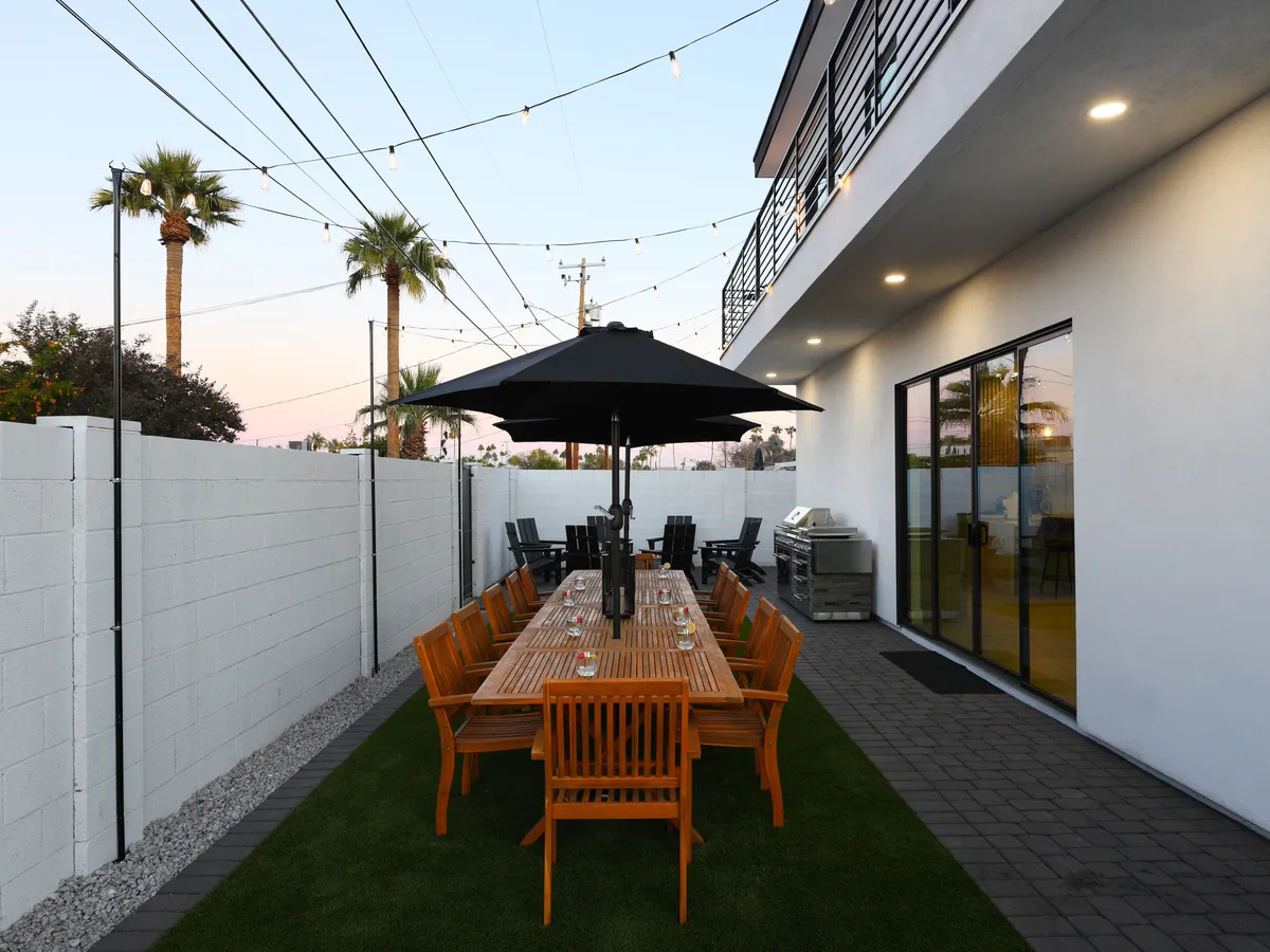 An exterior shot featuring a long wooden dining table under string lights, a black umbrella, a grill area, and palm trees in the background, all set up for al fresco dining and group gatherings.