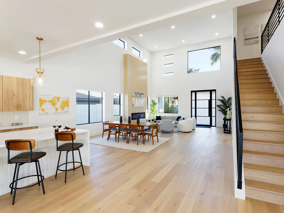 A stylish, open-plan space that showcases a dining table adjacent to a well-equipped kitchen island, a staircase, and a sunlit seating area. Large windows invite natural light, highlighting the blend of functionality and sophistication throughout the home.