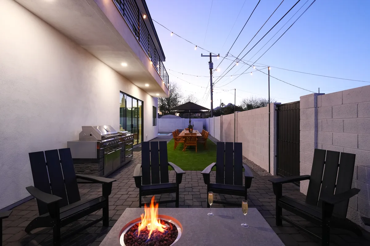 A cozy corner of the backyard showing a fire pit surrounded by comfortable outdoor seating, with a dining table and grill nearby. The space is illuminated by the soft glow of twilight, creating an inviting atmosphere for evening gatherings and sharing stories under the stars.