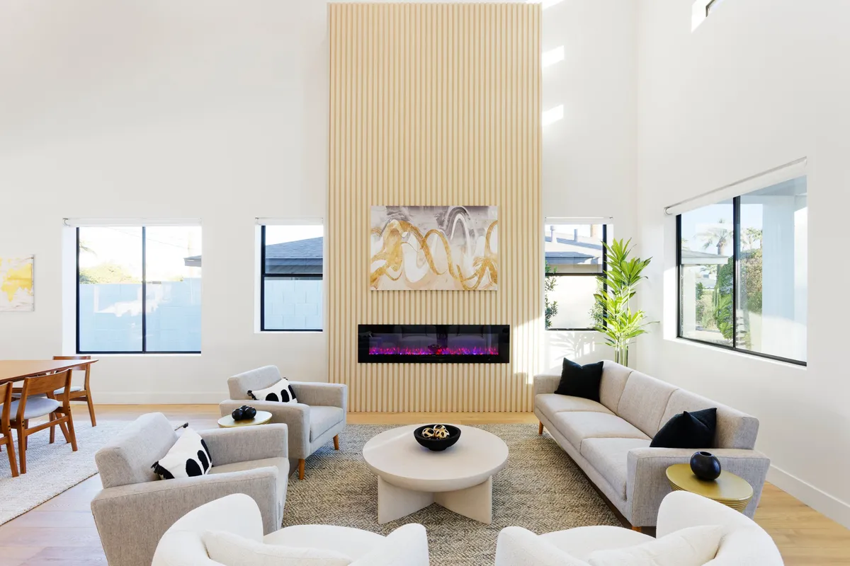 A sophisticated and welcoming open-plan living area featuring a modern fireplace with colorful lighting, comfortable designer sofas, striking artwork, and a glimpse of a dining table nearby. The atmosphere is set for relaxation and social gatherings, with the fireplace providing a cozy central anchor.